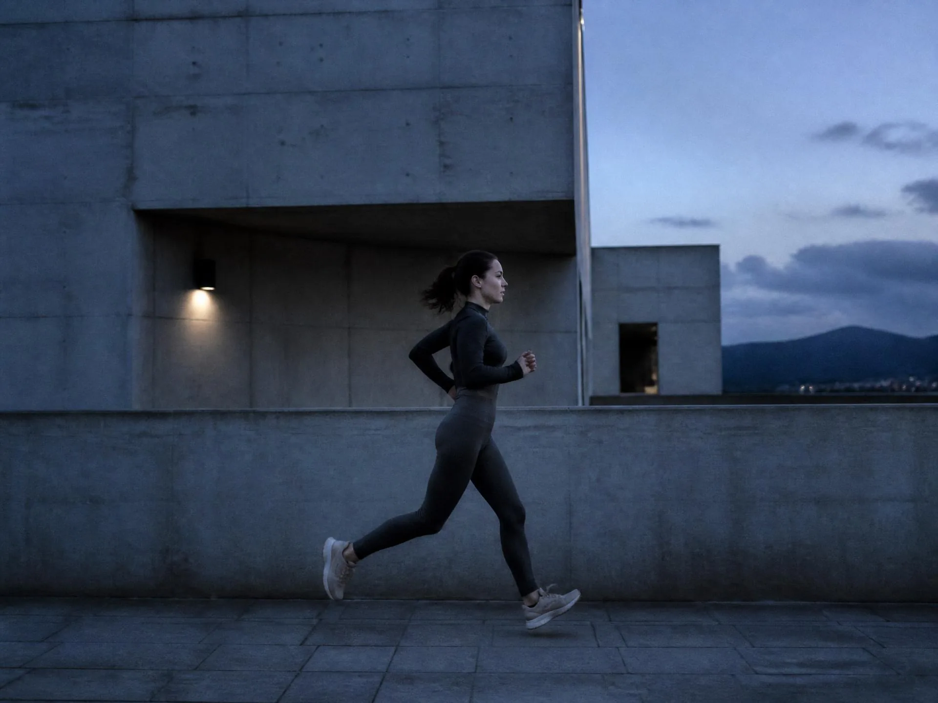 Runner in matte graphite activewear passing a concrete wall at blue hour